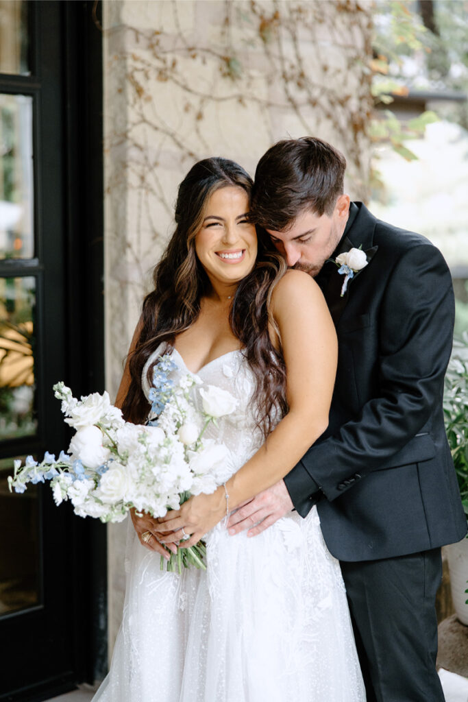 A bride in a white dress holding a bouquet smiles while the groom in a black suit hugs her from behind and kisses her shoulder.