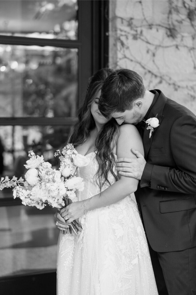 A bride holding a bouquet stands beside a groom who leans in to kiss her shoulder; both are dressed in formal wedding attire.