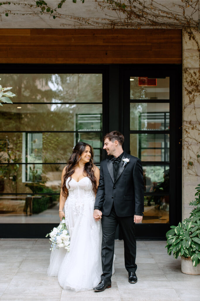A bride in a white gown and a groom in a black suit stand holding hands and smiling at each other outside a modern building.