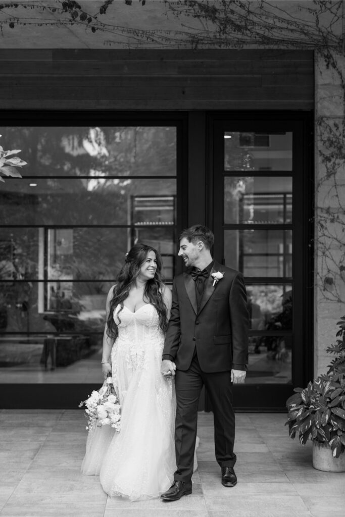 A bride and groom stand holding hands outside a building, looking at each other and smiling. The bride holds a bouquet. The photo is in black and white.