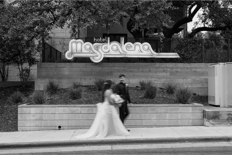 A bride and groom walk past a "hotel Magdalena" sign on a sidewalk, captured in black and white.