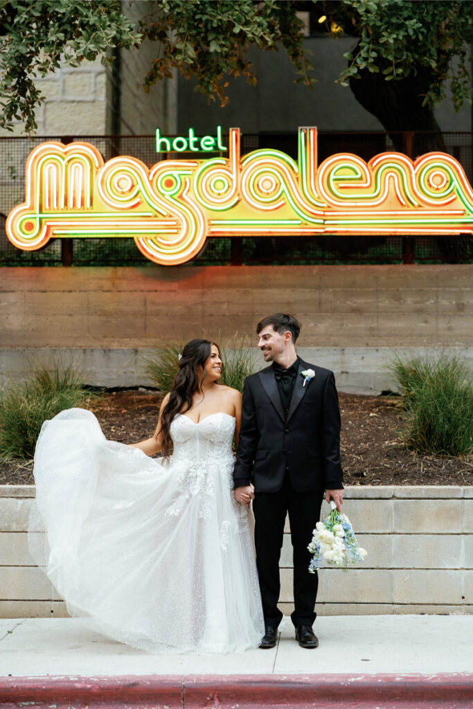 A bride and groom smile at each other while holding hands in front of a neon "hotel Magdalena" sign. The bride holds her dress and bouquet, and both are dressed formally.