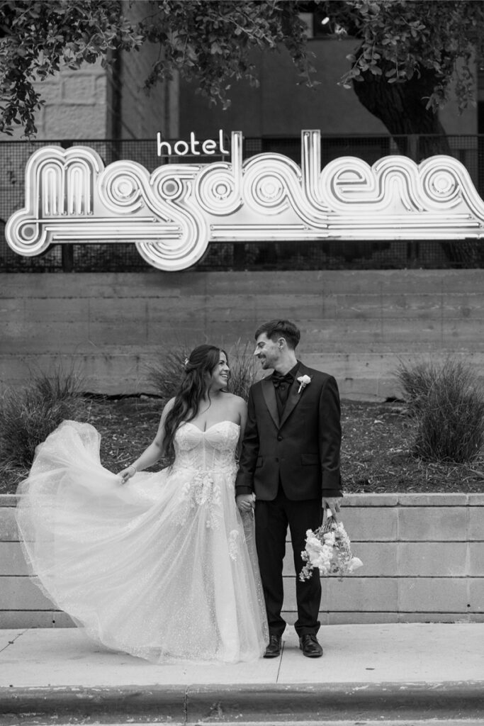 A bride and groom stand smiling in front of a neon "hotel Magdalena" sign; the bride holds her dress, and the groom holds a bouquet.