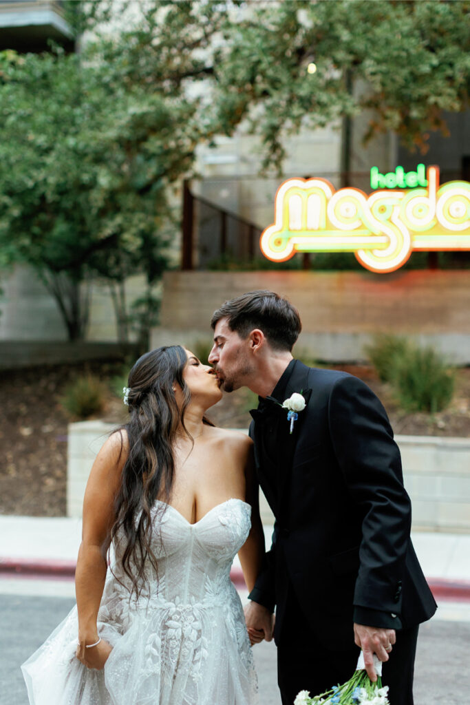 A bride and groom share a kiss outside, standing in front of a neon "hotel Magdalena" sign, both dressed in formal wedding attire.
