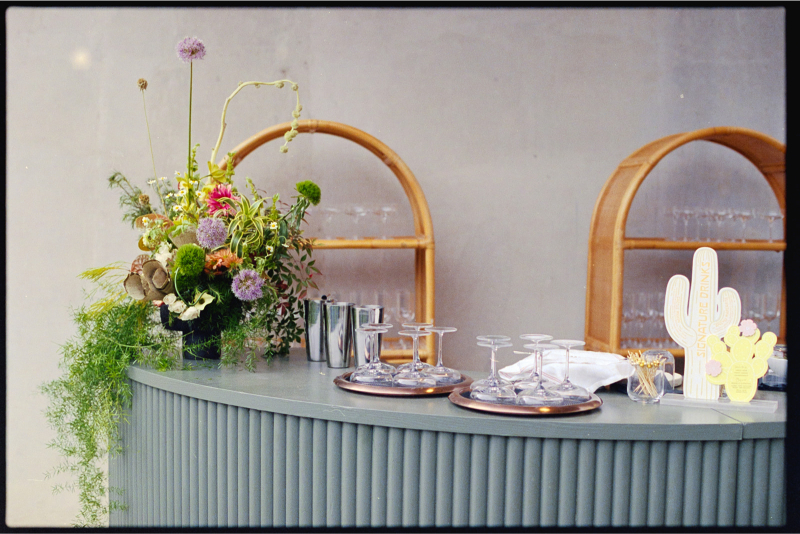 A modern bar counter with glassware, napkins, a floral arrangement, and decorative items in front of shelves holding glassware.