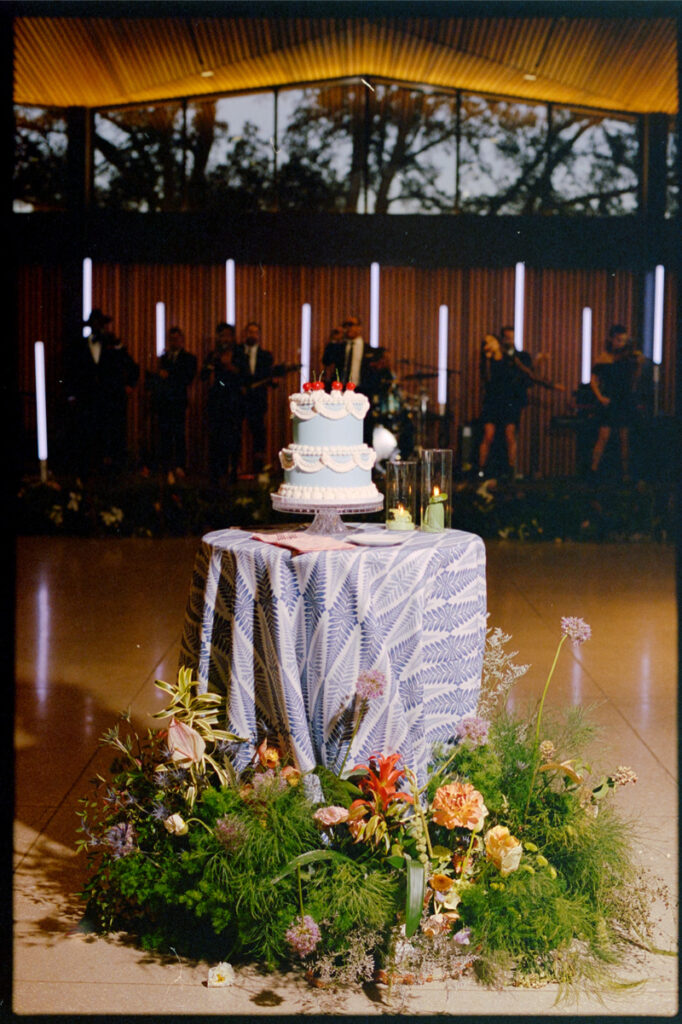 A two-tiered cake on a patterned tablecloth, surrounded by flowers, with a band playing in the background at an indoor event.