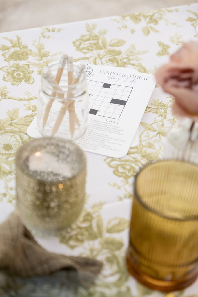 A floral tablecloth with a crossword puzzle card labeled “Anne & Drew,” a jar with wooden picks, a glittery candle holder, a ribbed amber glass, and a light pink flower.