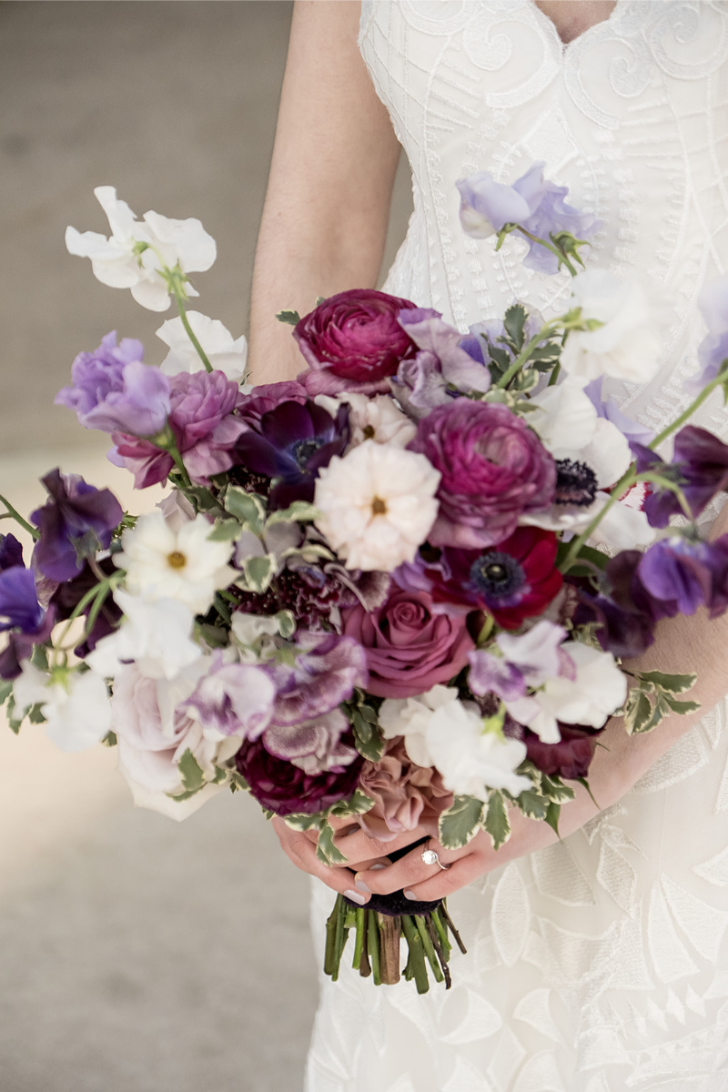 A person in a white dress holds a bouquet of purple, white, and pink flowers with greenery.