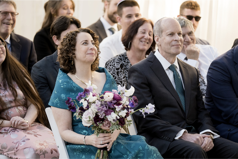 A woman in a teal dress holds a bouquet of flowers and sits beside a man in a suit, surrounded by other seated guests at an event.