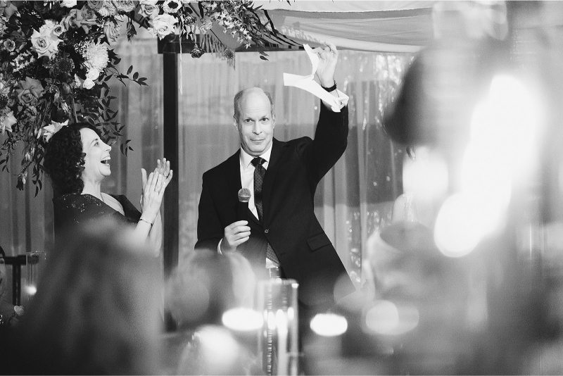 A man in a suit holds up a piece of paper while speaking into a microphone, as a woman beside him claps, surrounded by flowers and blurred lights.