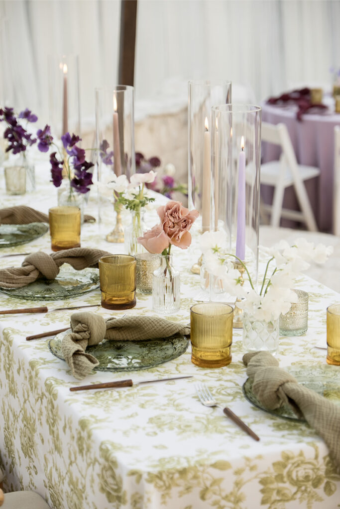 Elegant table setting with green floral tablecloth, amber glasses, green plates, beige napkins, and assorted flowers in vases. Tall glass candle holders with purple candles add height.