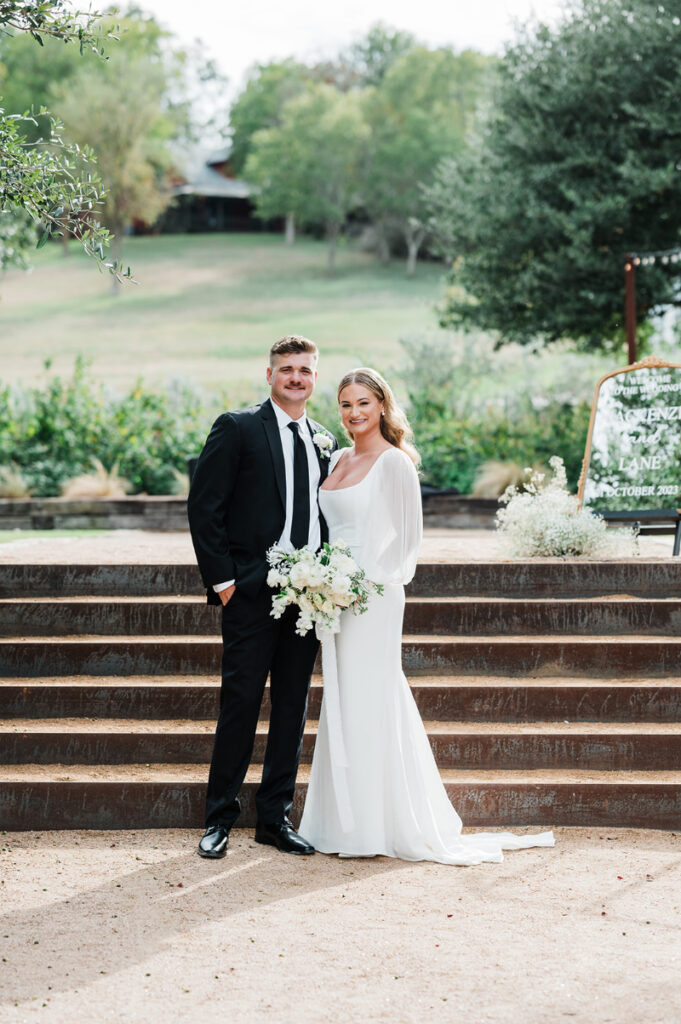 A bride in a white dress and a groom in a black suit stand together outdoors, smiling in front of stone steps with greenery in the background.