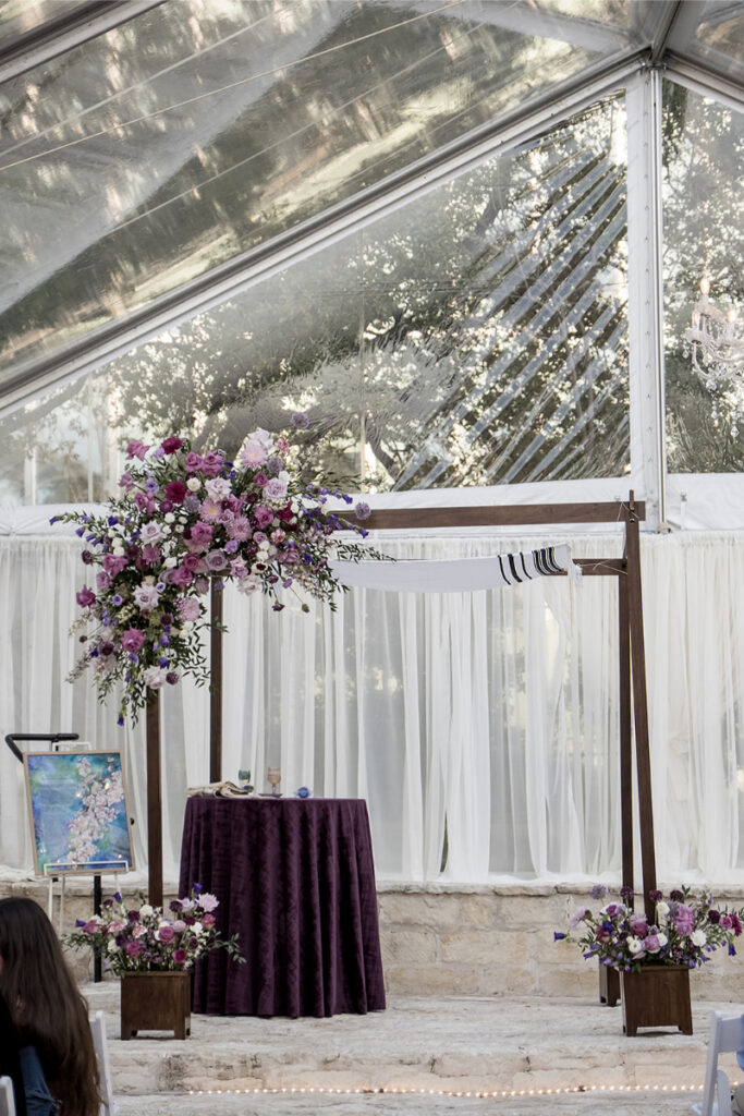 A wedding chuppah in a wedding venue in Austin is decorated with purple flowers stands under a clear tent. A table with a purple ta