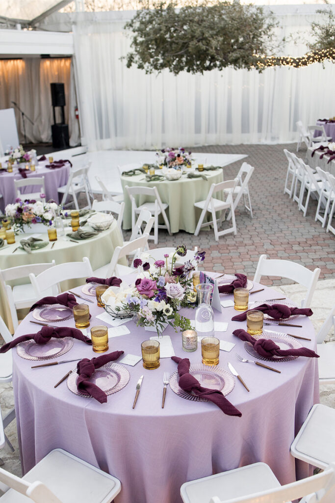 Round tables with lavender and light green tablecloths are set for an event with floral centerpieces, gold glasses, and white chairs in an outdoor covered area.