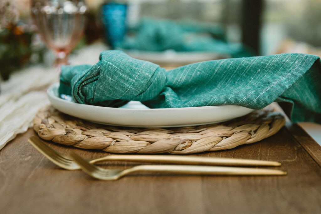 A close-up of a table setting by features a white plate, green cloth napkin, woven placemat, and gold-colored fork and knife on a wooden table.