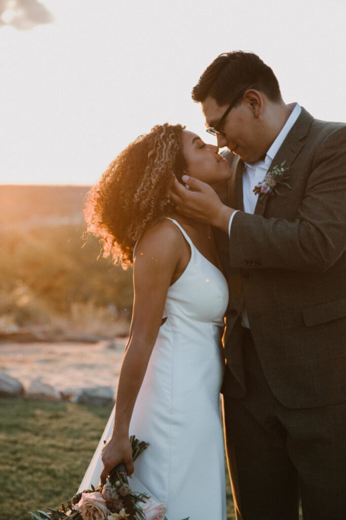 A couple in wedding attire stands outdoors at sunset, the groom gently holding the bride's face as they prepare to kiss. The bride holds a bouquet of flowers—an unforgettable moment.