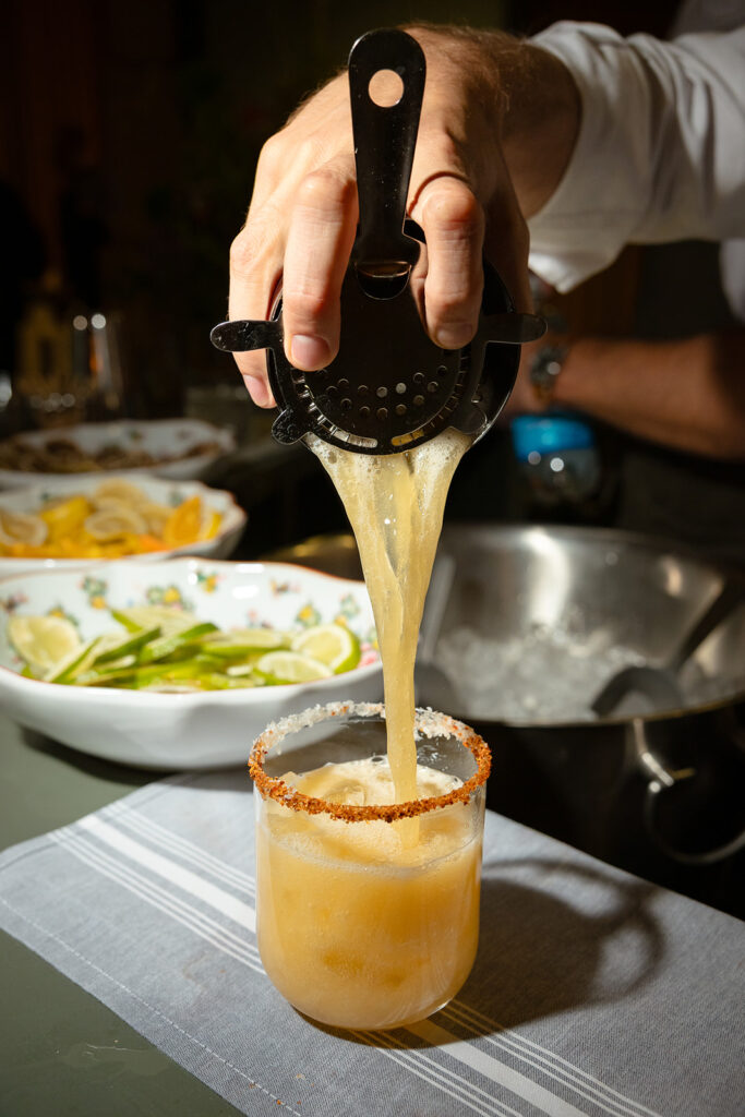 A person pours a yellow cocktail through a strainer into a glass with a chili-salt rim, with sliced fruit artfully arranged in the background.