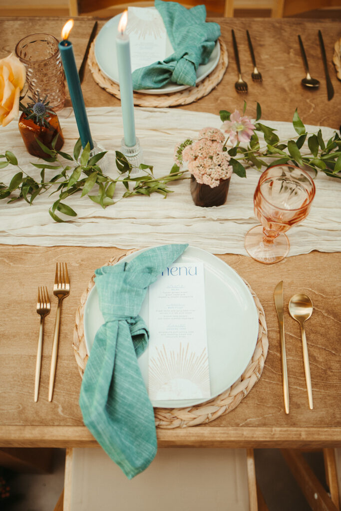 A wooden table set with a white plate, gold cutlery, a blue napkin, a pink glass, a menu, blue candles, small flowers, and a leafy table runner styled by a Jackson Hole wedding planner.