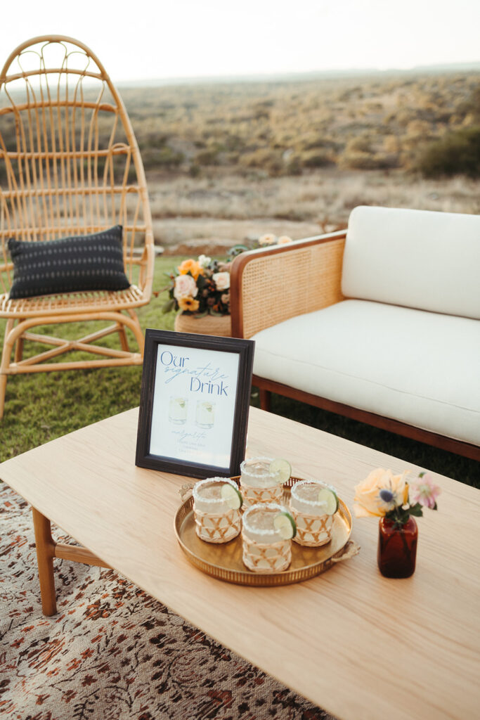 A wooden table with a tray of four cocktails, a small flower vase, and a framed drink menu sits in front of a wicker chair and sofa—an inviting outdoor setup styled by a Jackson Hole wedding planner.