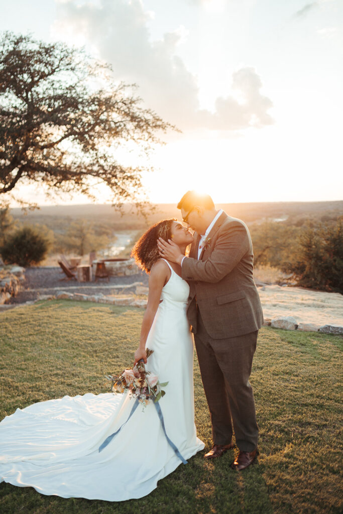 A bride and groom share a kiss outdoors at sunset, with the bride holding a bouquet and wearing a white dress, and the groom in a suit.