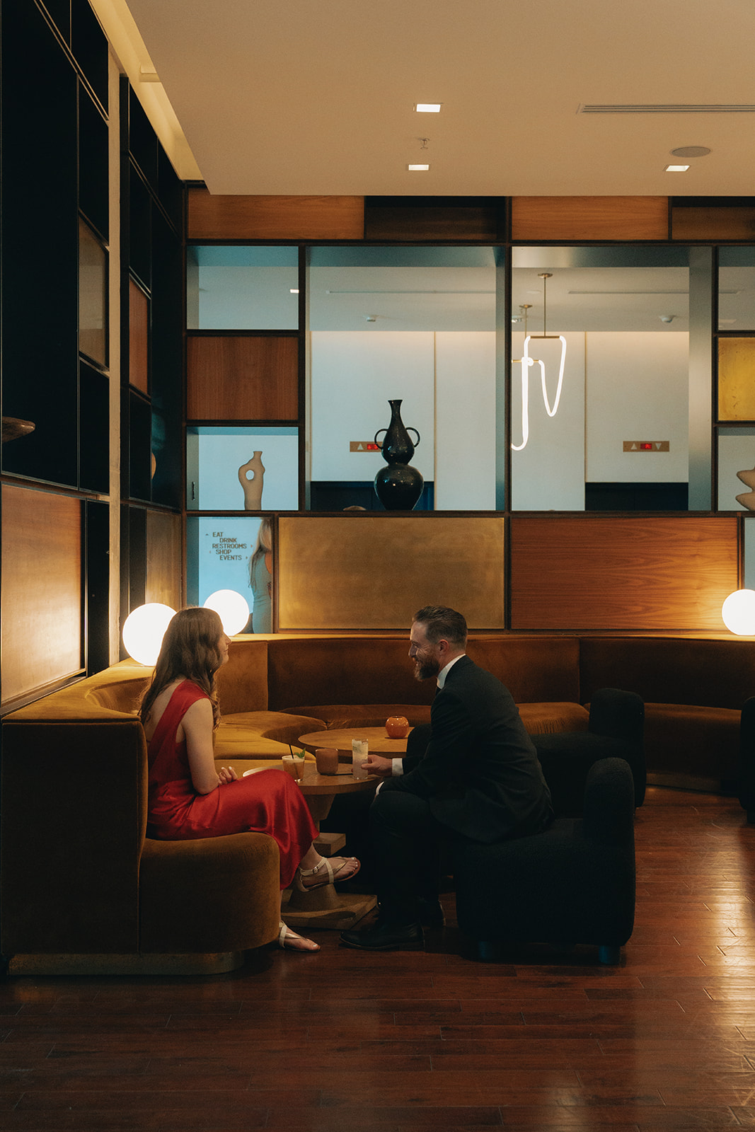 A man and woman sit across from each other at a table in a modern, dimly lit lounge, engaged in conversation with drinks in front of them.
