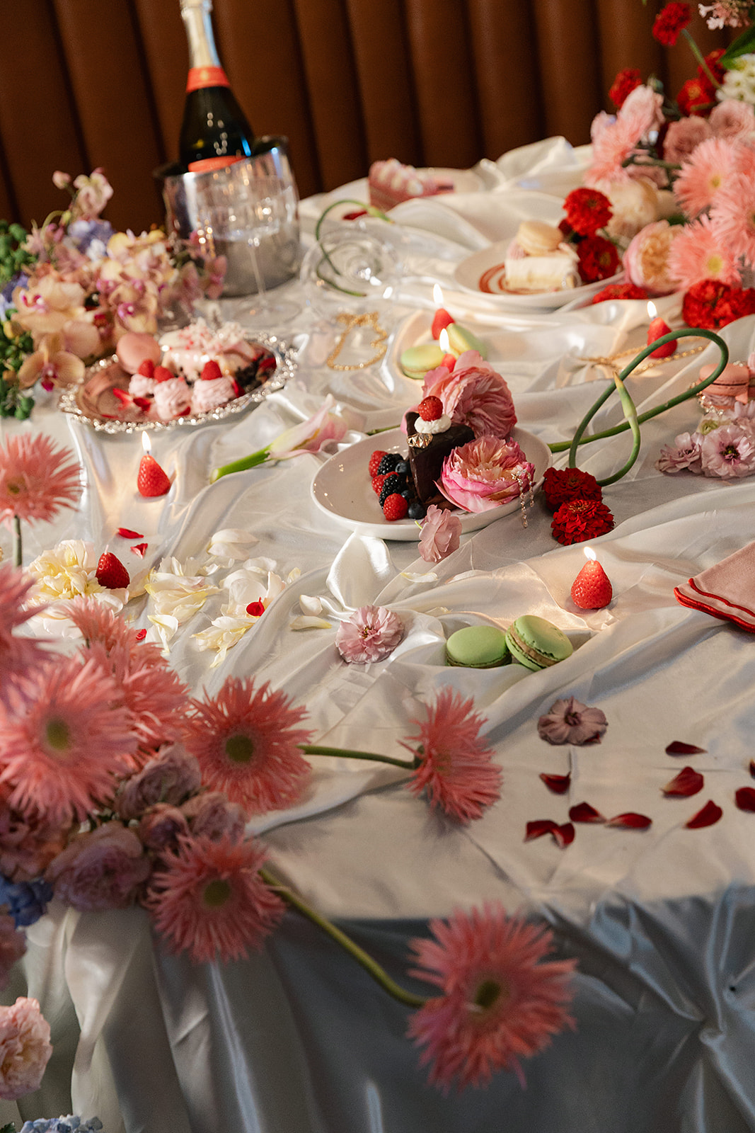 A table is decorated with pink flowers, macarons, strawberries, desserts, rose petals, and a bottle of champagne in an ice bucket on a white tablecloth.