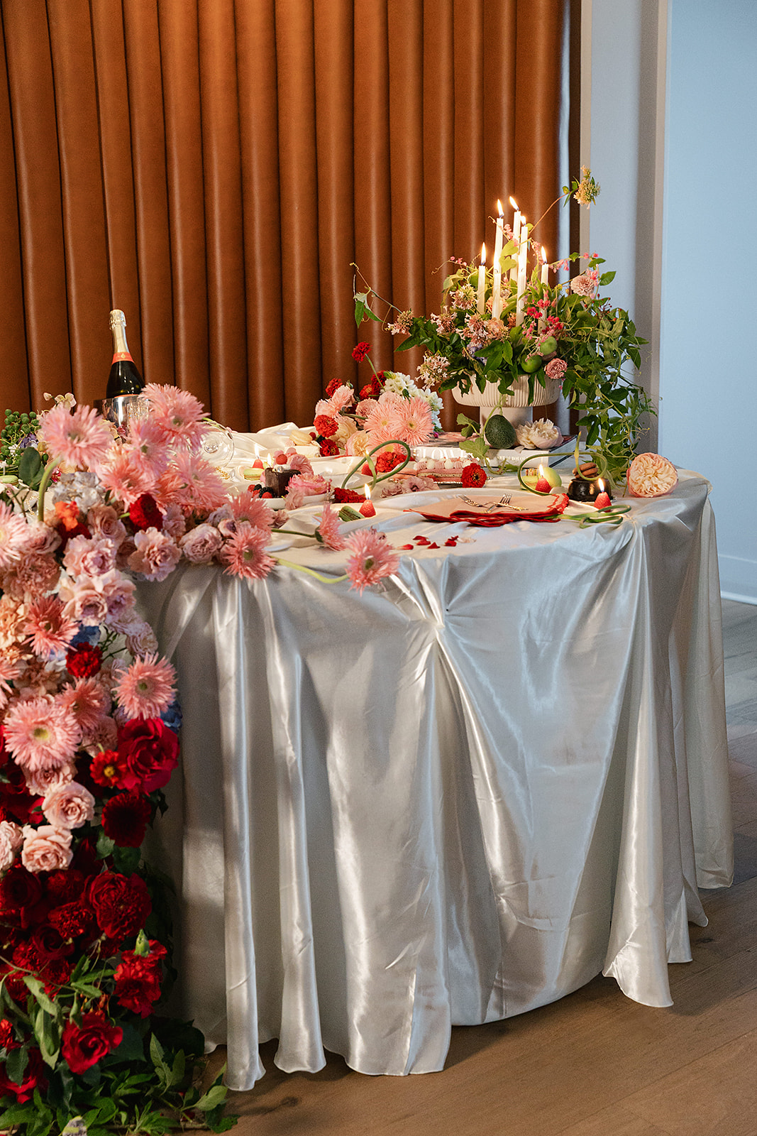 A round table with a white satin tablecloth is set with flowers, candles, pomegranates, grapes, and a bottle of champagne against a backdrop of brown curtains.