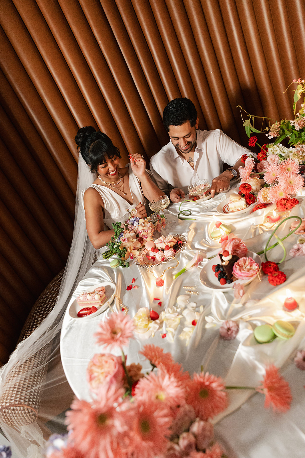 A bride and groom sit at a flower-decorated table, enjoying dessert together, surrounded by pink and red floral arrangements and assorted sweets.