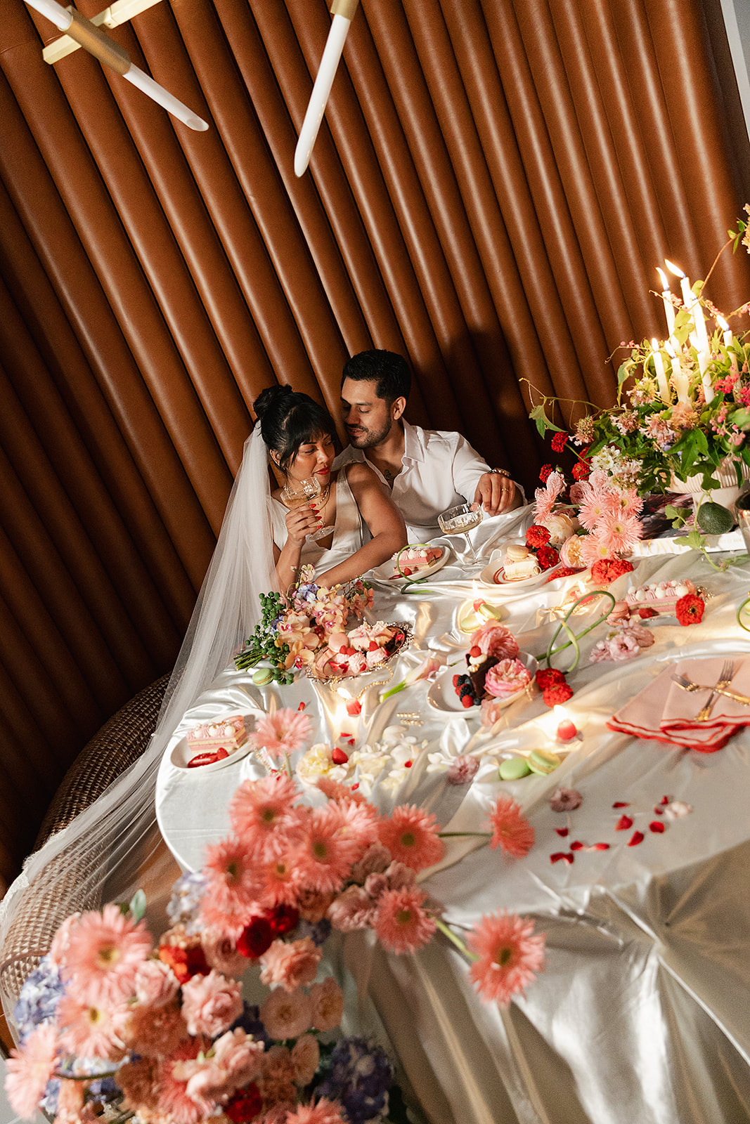 A bride and groom sit closely at a decorated table with flowers, candles, and food, sharing a romantic moment in an elegant indoor setting.
