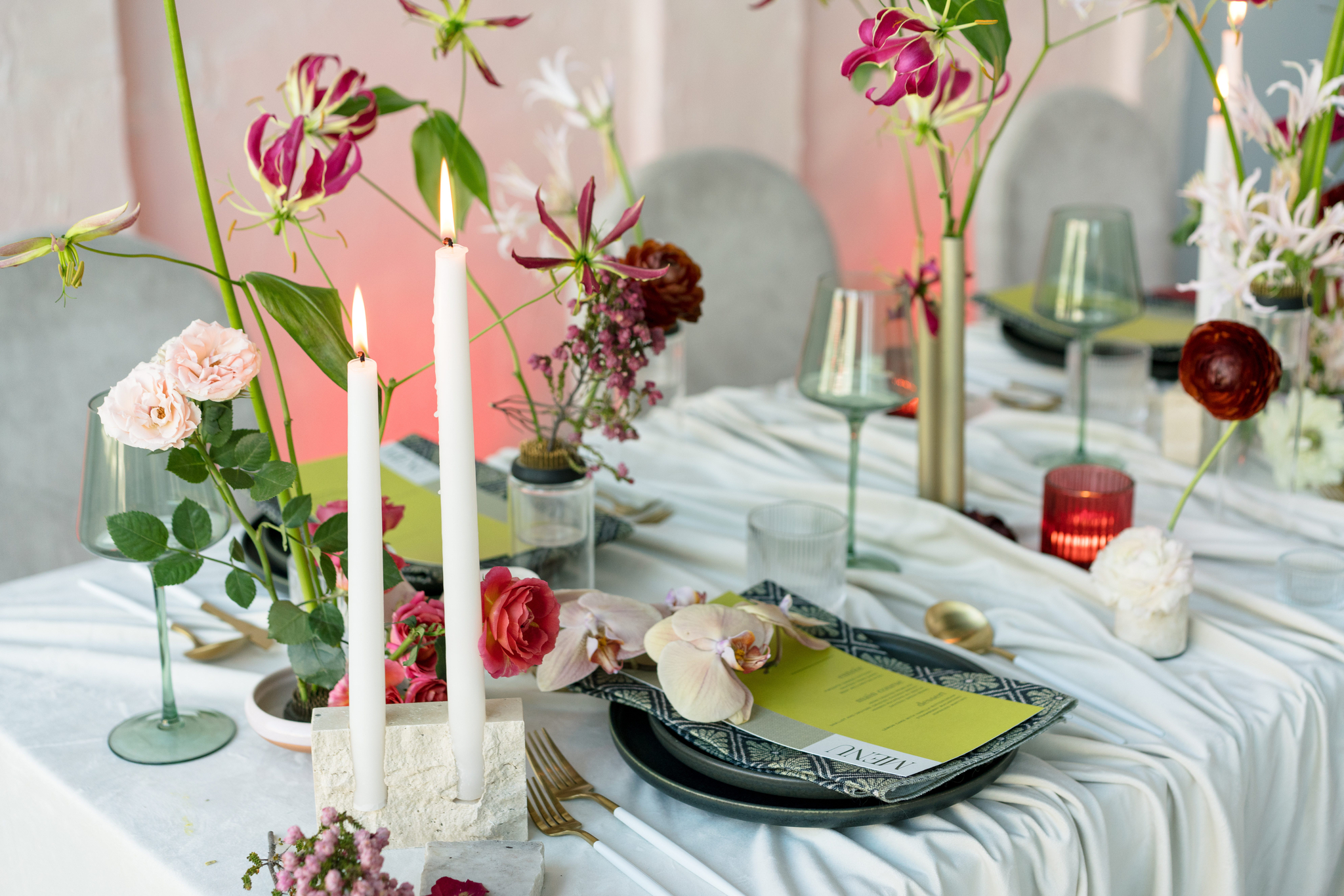 A table set for a formal event with white tablecloth, floral arrangements, two lit candles, glassware, plates, menus, and gold cutlery.