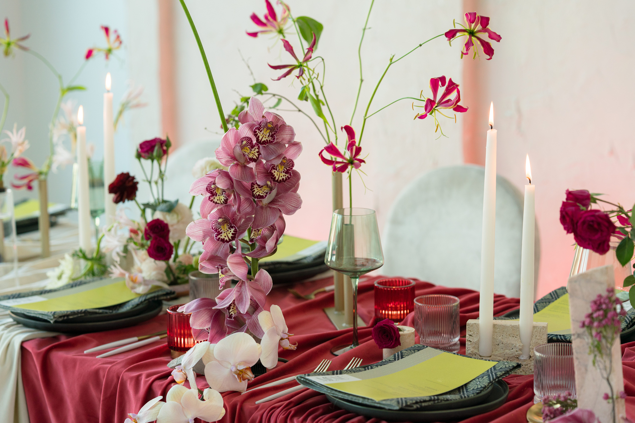 A formal dining table with red and white floral arrangements, green napkins, tall white candles, red glassware, and plates set on a red tablecloth, a perfect representation of a messy luxury wedding design.