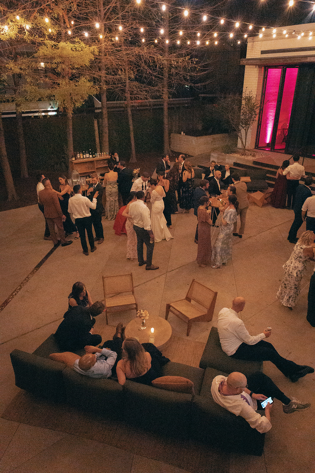 A group of people dressed in formal attire gather and socialize on an outdoor patio at night, with string lights overhead and seating areas scattered around.