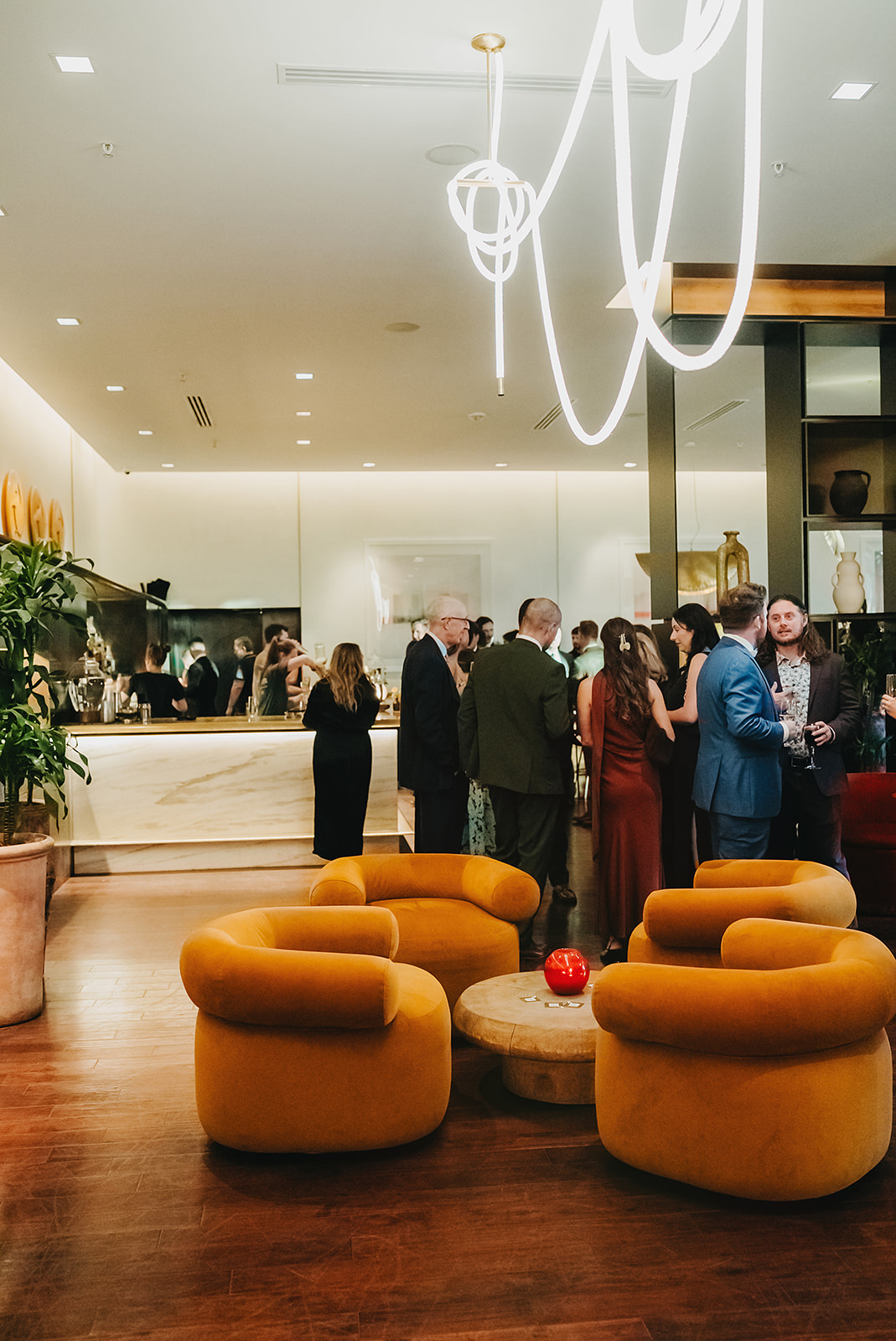 People gather and socialize in a modern lounge with yellow chairs, wooden floors, a marble bar, and contemporary lighting.