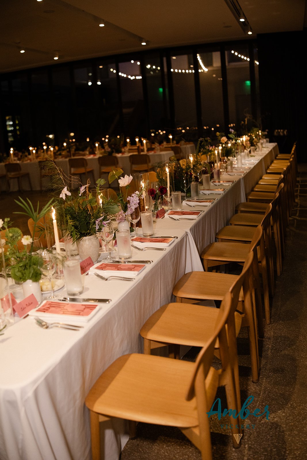 A long dining table is set with plates, glasses, menus, name cards, candles, flowers, and wooden chairs in a dimly lit room, a messy luxury wedding design.