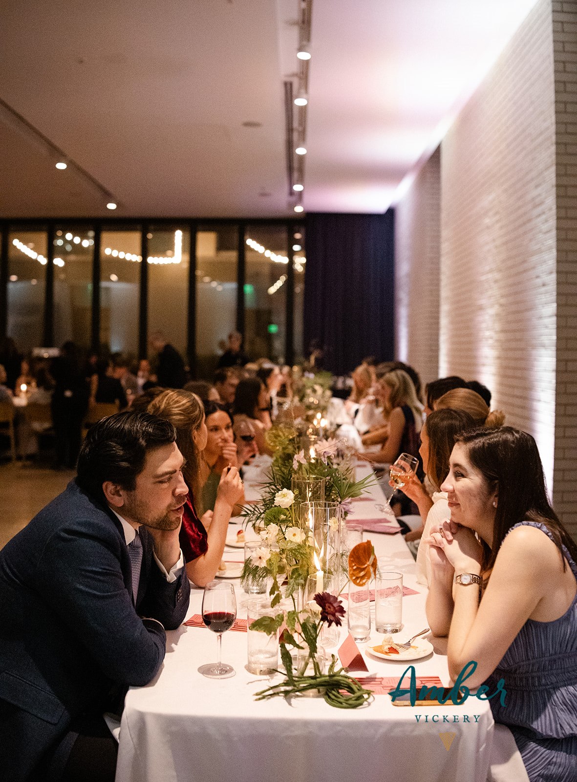 Guests sit at a long, decorated table in a softly lit indoor venue, engaged in conversation and surrounded by candles and flowers.