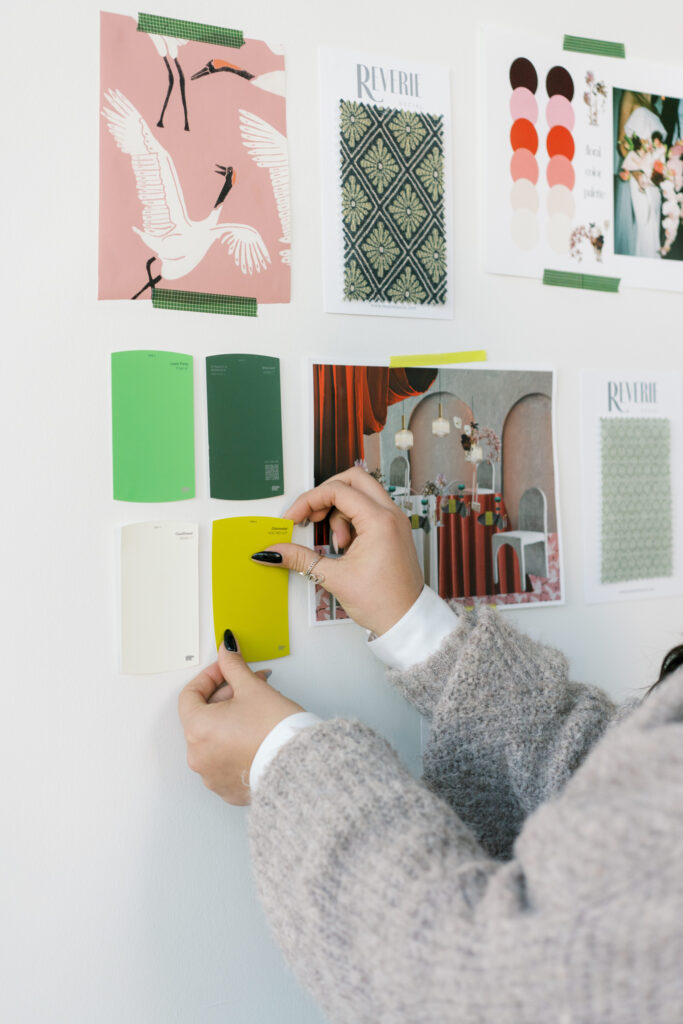 Sydney, California wedding planner and owner of In Ink Weddings, holds paint swatches against a white wall displaying design inspirations, color palettes, and patterned images taped up for reference.