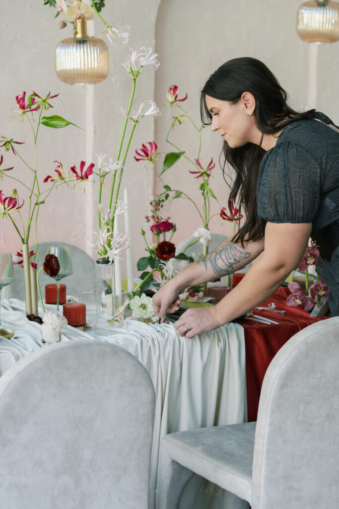 Sydney from In Ink Weddings, a California wedding planner, arranges flowers on an elegantly set dining table with white and red accents, surrounded by floral centerpieces and neutral-colored chairs.