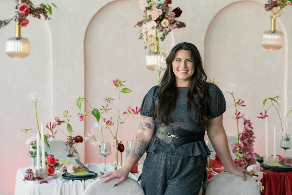 Sydney from In Ink Weddings, Austin-based and California wedding planner, stands in front of an elegantly set table with flowers, candles, and hanging lights.