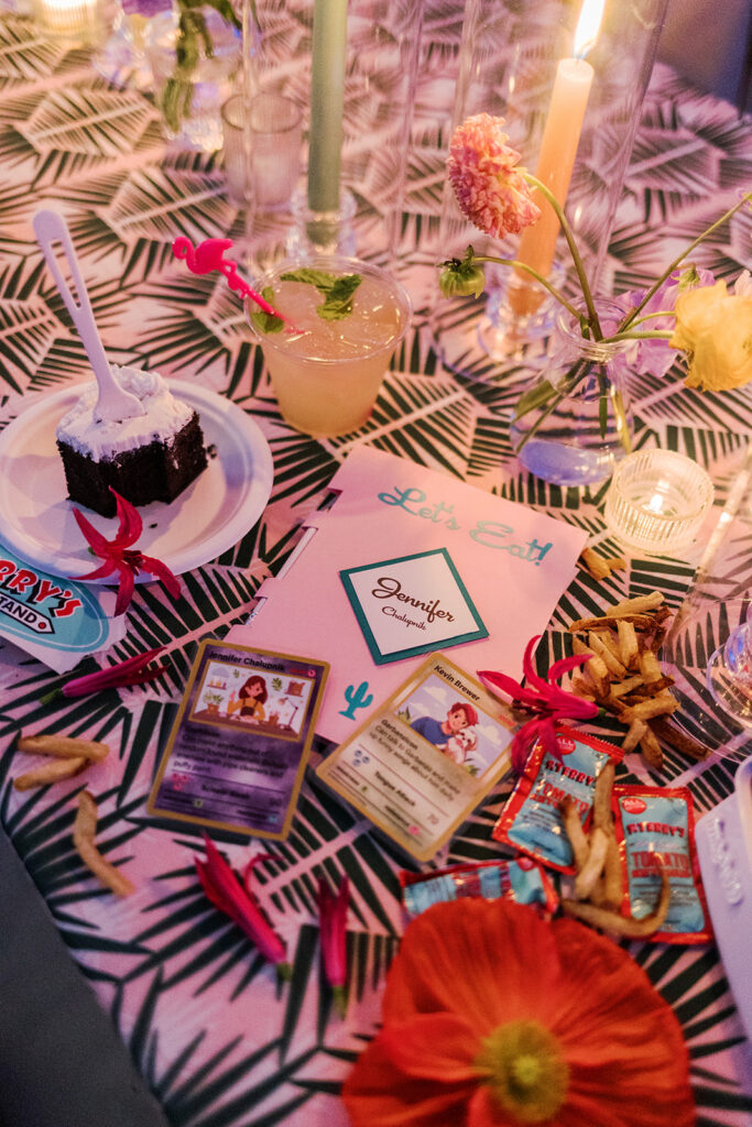 A decorated table with a patterned cloth features a slice of cake, a cocktail, a menu labeled “Jennifer,” Pokémon cards, fries, flowers, and various snacks under candlelight.