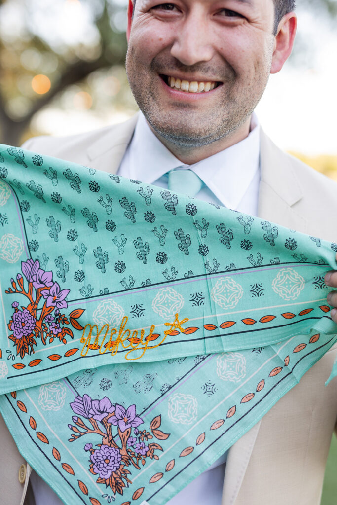 A man in a light suit holds a turquoise bandana with cactus patterns, purple flowers, and the word "Mikey's" embroidered in yellow.