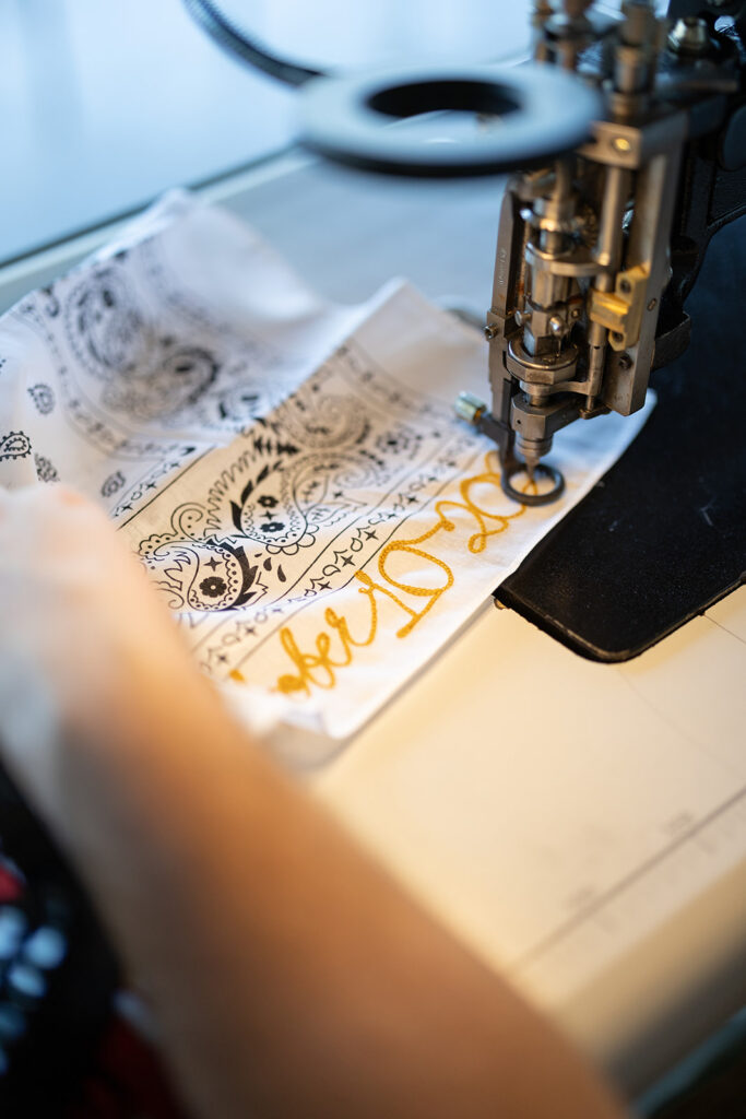 A sewing machine embroidering gold thread onto a white fabric with black patterned designs, with a person's hand guiding the material.