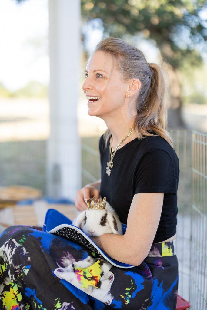 A woman wearing a black shirt and colorful skirt sits outside, smiling, while holding a rabbit with a small crown on its head.