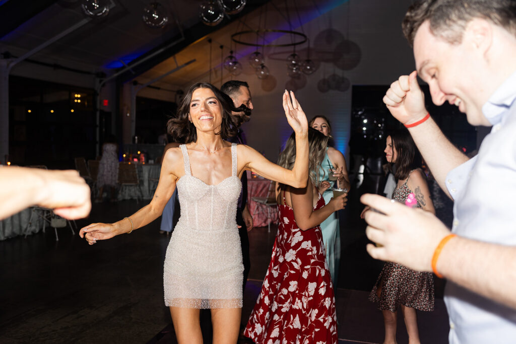 A group of people dance and celebrate indoors under hanging lights. The woman in front wears a sparkly dress and smiles with her hands raised.