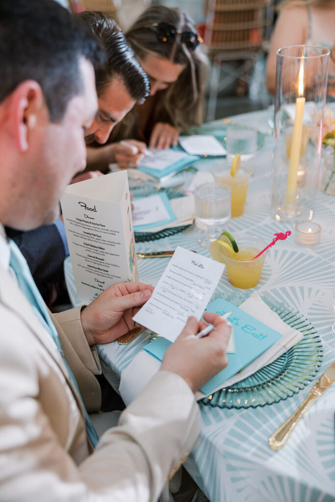 A wedding weekend guest sits at a decorated table and reads a printed note while holding a menu; drinks, candles, and place settings are visible.