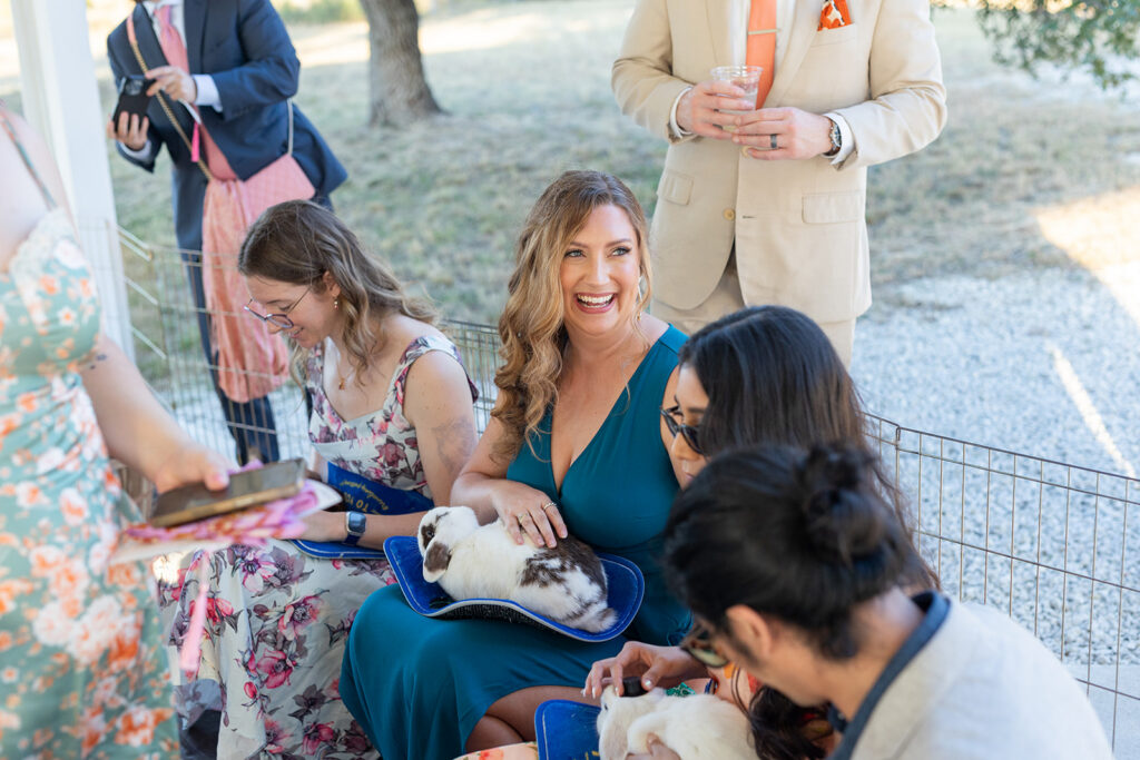 Wedding guests sit together outdoors, some holding rabbits on their laps and smiling, near a metal pen, as they take in all the activities for the couple's wedding weekend.