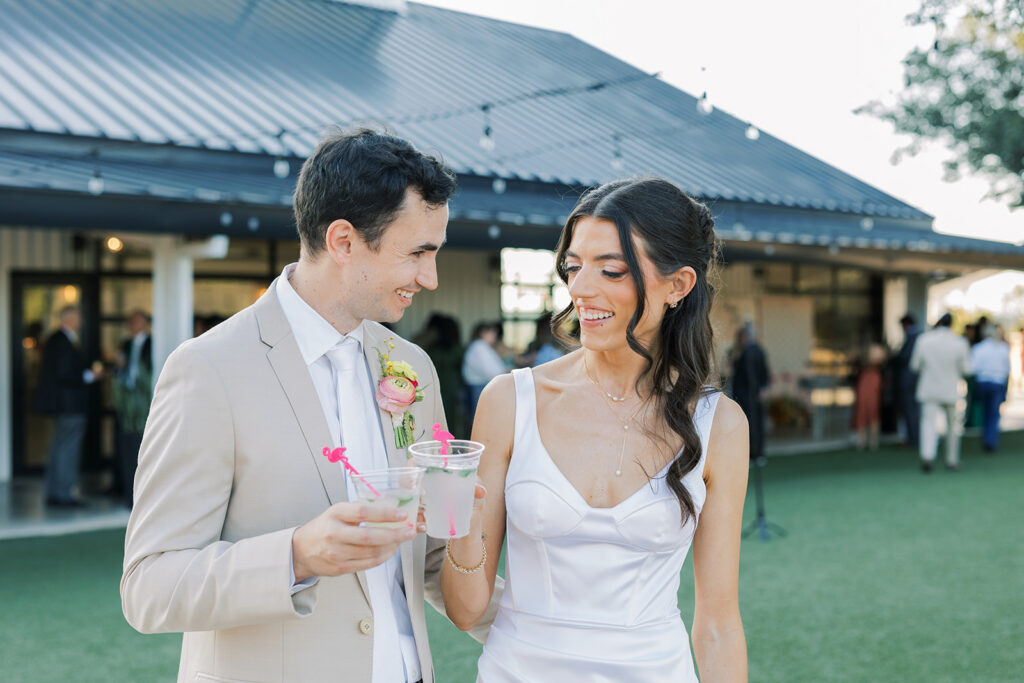 A man in a tan suit and a woman in a white dress holding drinks and smiling at each other outdoors, with people and a modern building in the background.