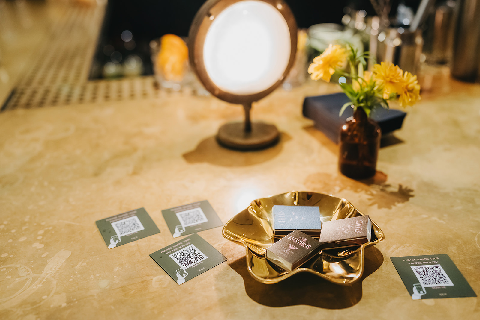 A gold dish with business cards sits on a marble countertop near scattered QR code cards, a small lamp, and a brown vase with yellow flowers.