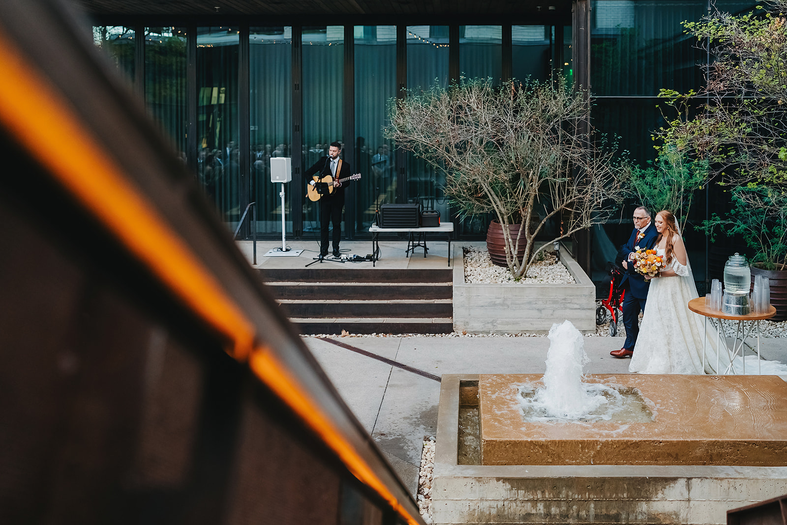 A bride holding a bouquet stands with her father near a small fountain in the courtyard at South Congress Hotel while a musician with a guitar performs in the background, representing how sound create an elevated guest experience.