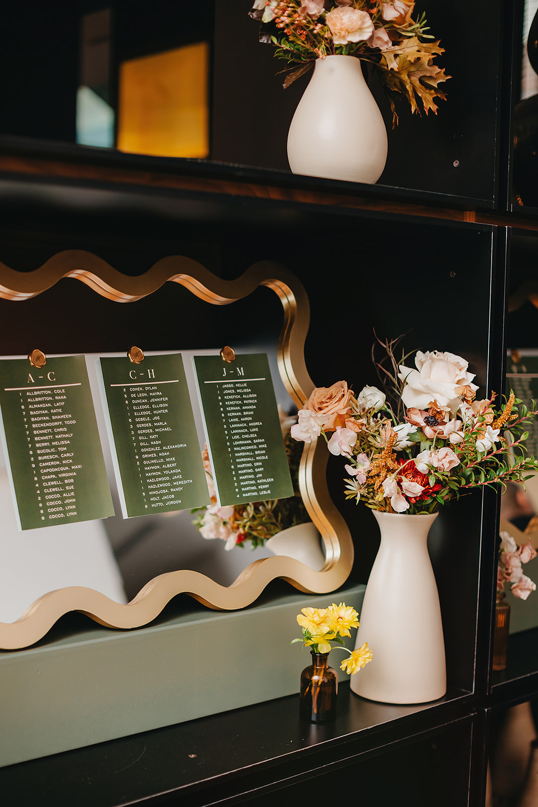 A decorative mirror displays three seating charts secured with gold pins. Two white vases with assorted flowers and a small vase with yellow flowers sit nearby on black shelves.