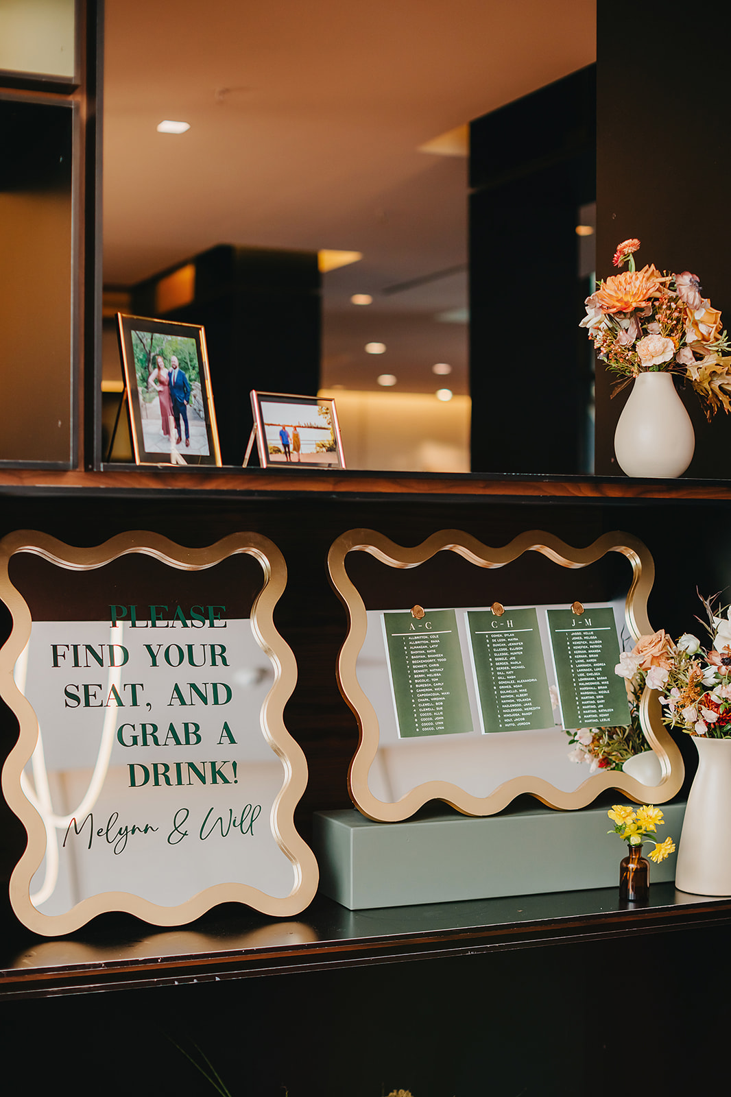 A wedding seating chart display with framed signs, including a message to find your seat and grab a drink, surrounded by flowers and photos.
